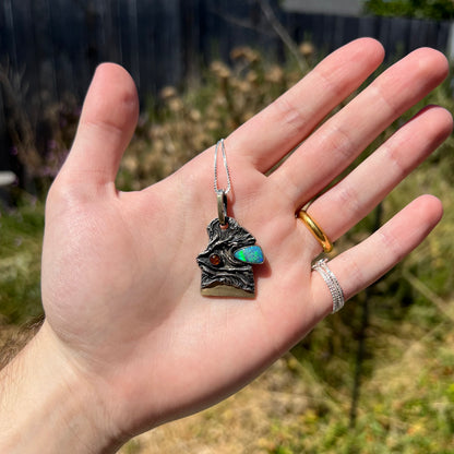 Hand holding a brutalist men's Australian opal doublet pendant in sterling silver under natural outdoor light against a blurred green backdrop.
