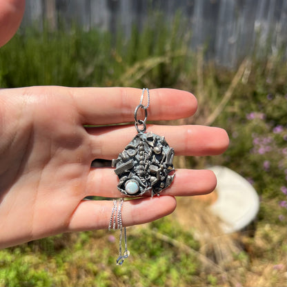 A brutalist opal necklace made from sterling silver bench scrap, held in a hand at a distance in sunlight.
