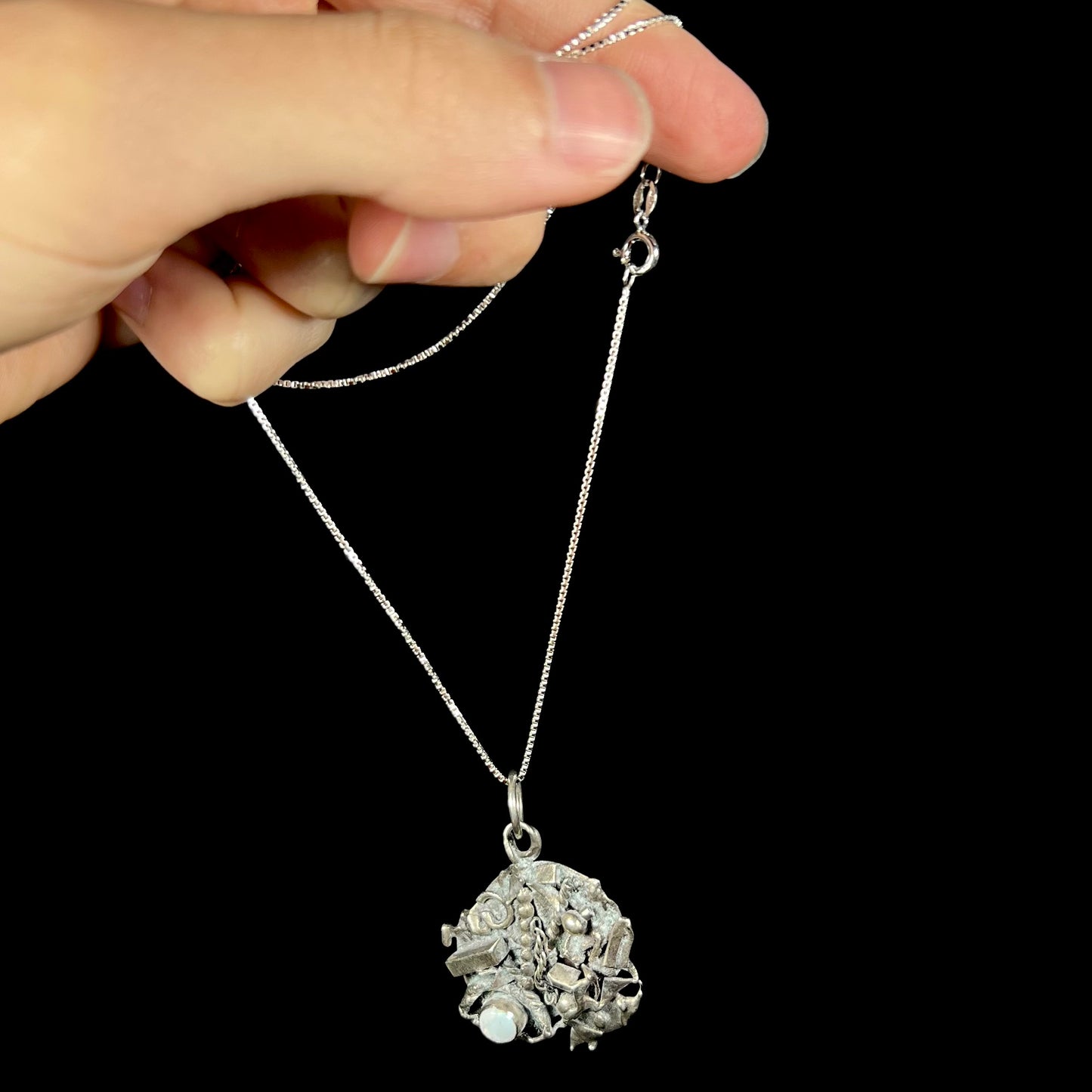 A brutalist silver opal necklace, dangling by its chain from a hand against a black backdrop.