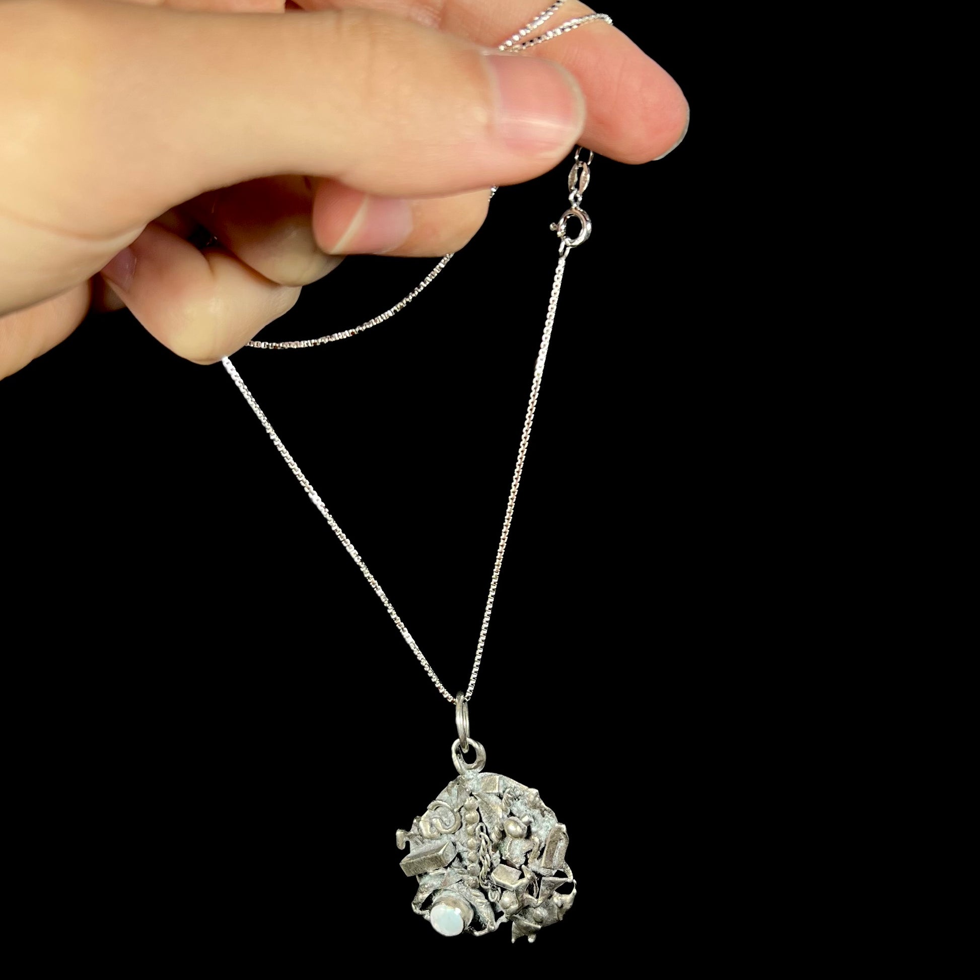 A brutalist silver opal necklace, dangling by its chain from a hand against a black backdrop.