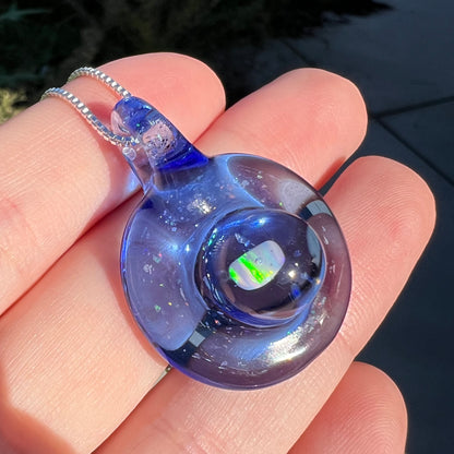 Angled sunlit close-up of a glass and opal pendant by artist, Cameron Scott, shown held in a hand against an outdoor concrete background.