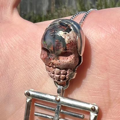 Close-up angled view of sterling silver and 14k gold-accented skeleton pendant set with carved cantera opal skull exhibiting needle-like black tourmaline inclusions, held in natural light