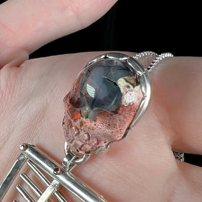 Black tourmaline needle inclusons seen in carved Mexican cantera opal skull viewed close-up in sterling silver skeleton pendant, held in hand