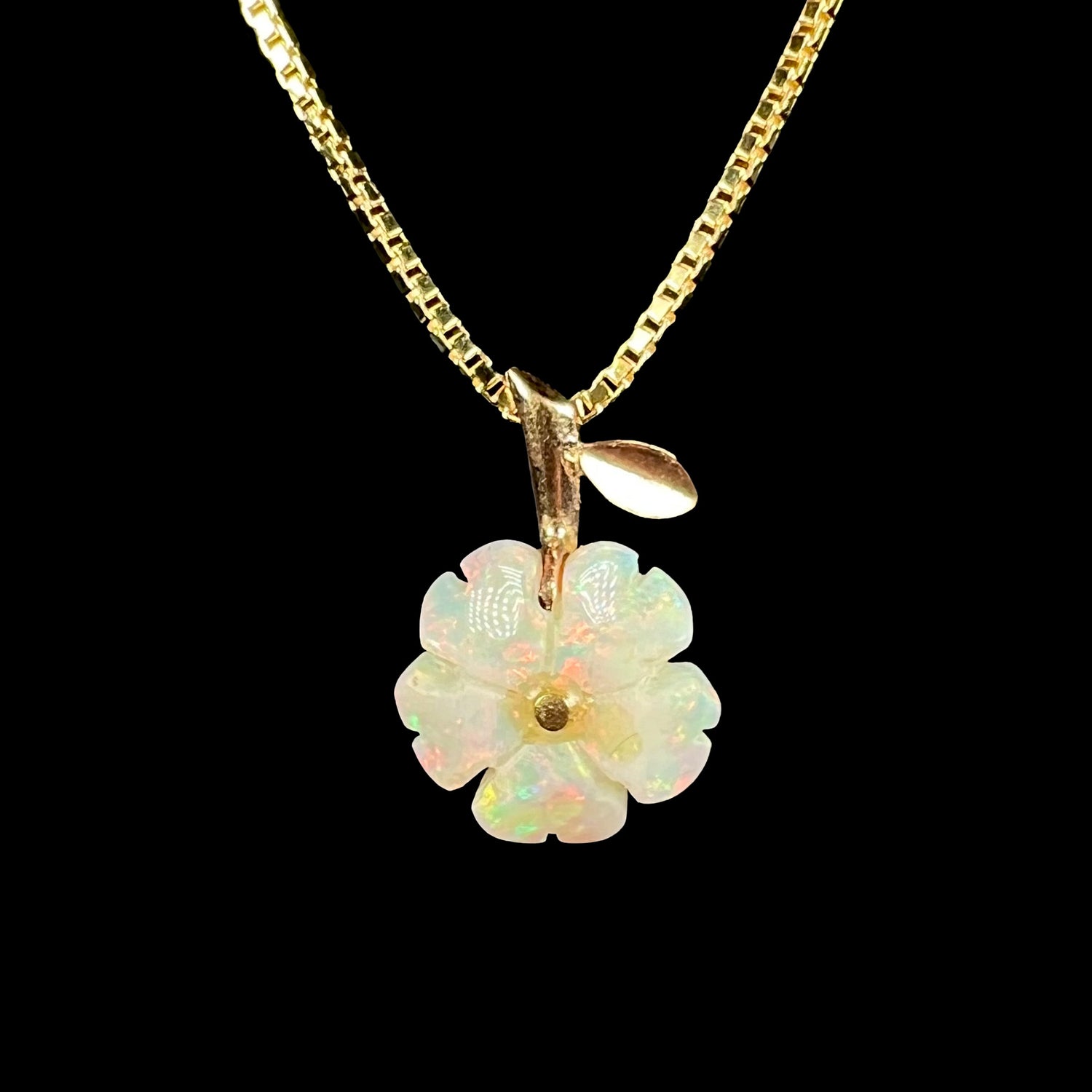 Carved white opal flower necklace with a 14k yellow gold stem-design bail by Parle on a gold-tone box chain against a black background.