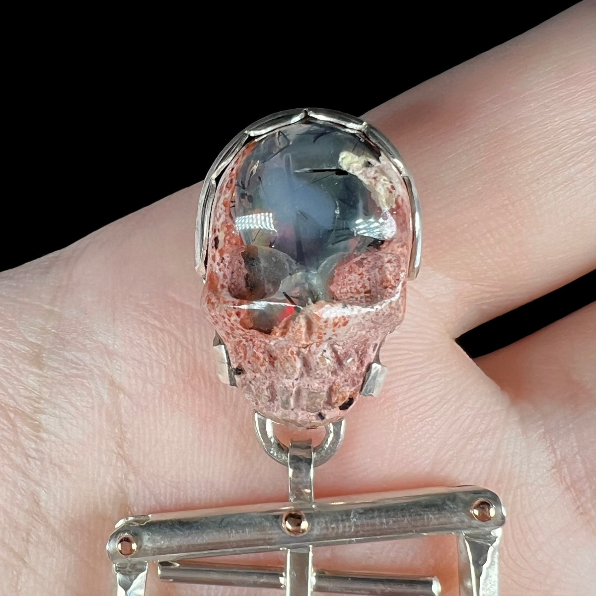 Close-up of carved Mexican cantera opal skull set into sterling silver skeleton pendant with three 14k yellow gold rivets, held in hand