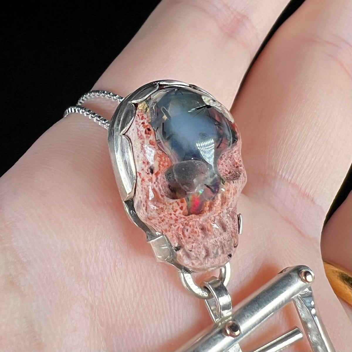 Angled close-up of red flash in carved cantera fire opal skull mounted in two-tone skeleton pendant, held in hand 