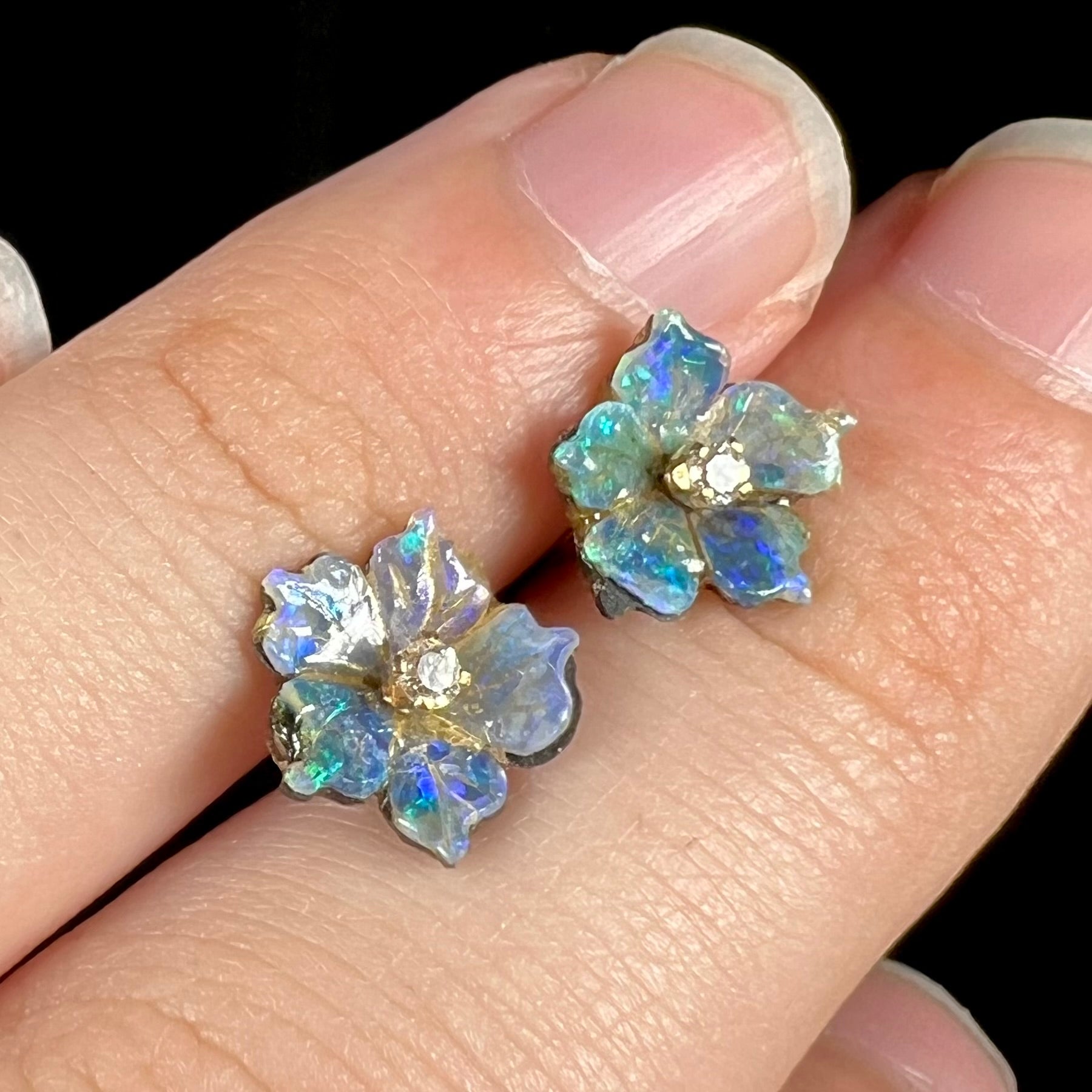 A pair of carved boulder opal doublet flower earring jackets with gold CZ studs shown from an angle against fingers of a hand.