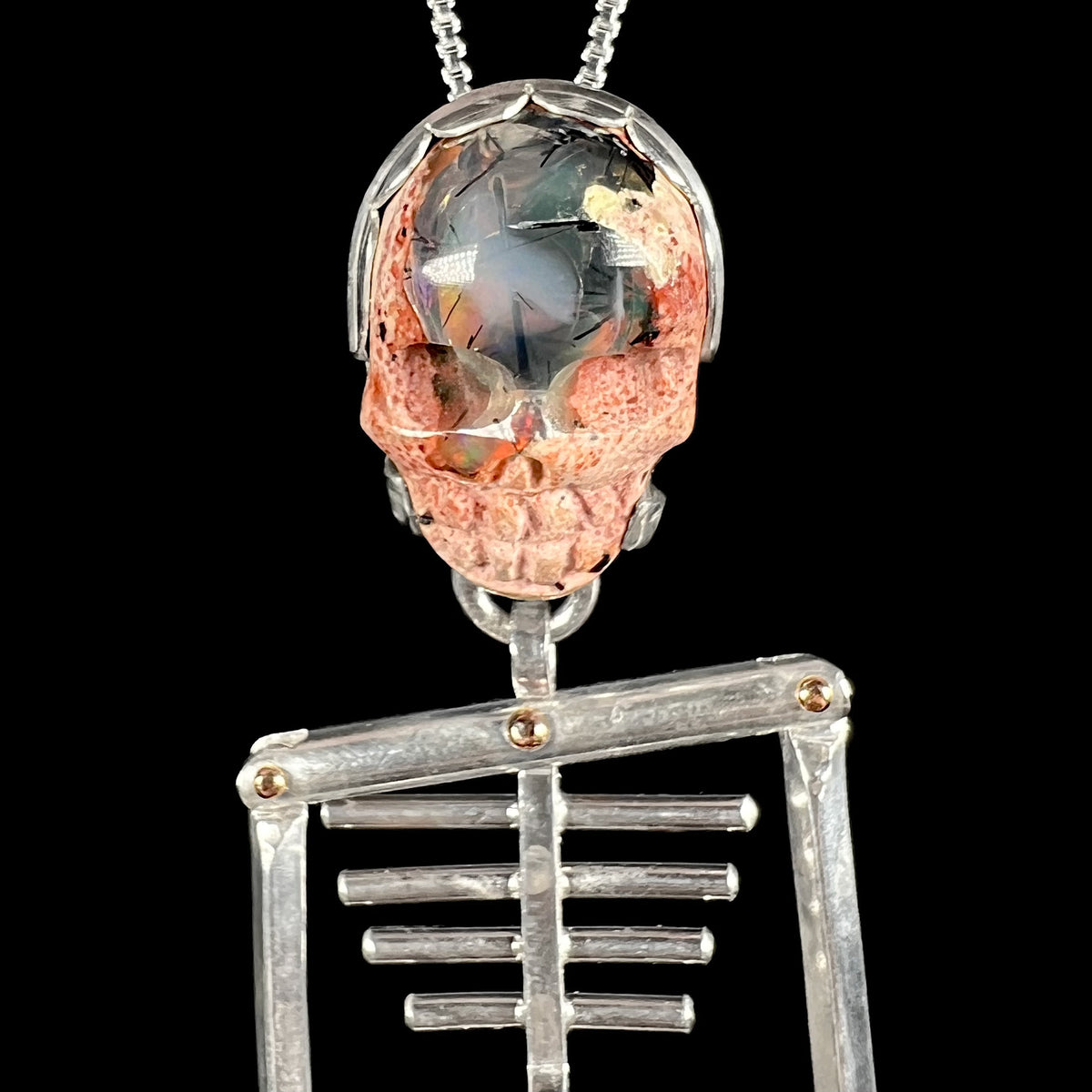 Close-up of carved Mexican cantera opal skull mounted in sterlnig silver and 14k gold-accented skeleton pendant.  The opal has black tourmaline needle inclusions