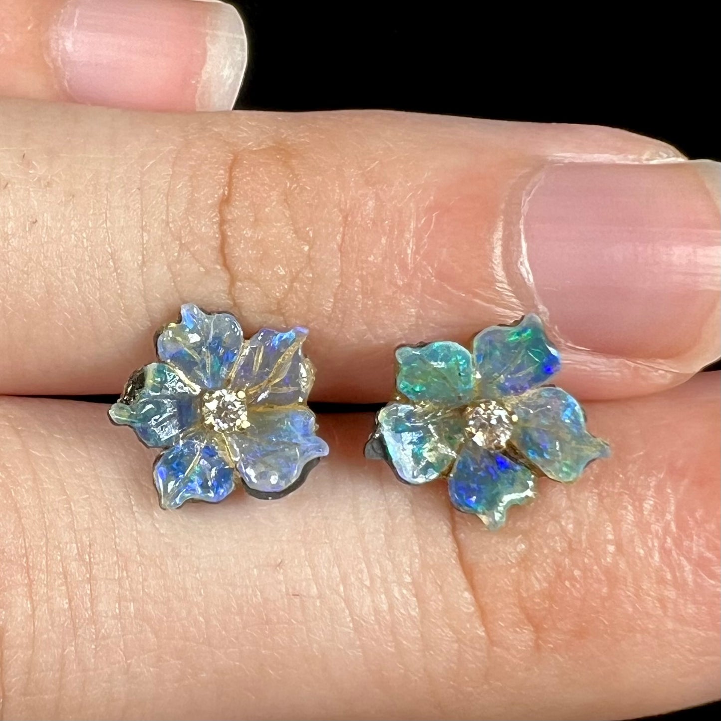 Close-up front view of a pair of carved boulder opal doublet flower earring jackets with gold CZ studs shown between fingers.