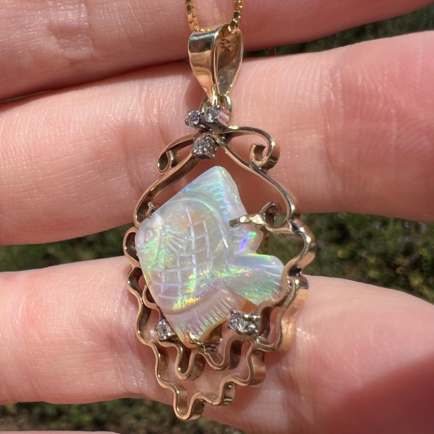 Angled close-up of carved opal fish pendant in 14k gold with diamond accents, held in sunlight