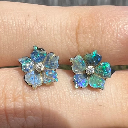 A pair of carved boulder opal doublet flower earring jackets with gold CZ studs shown close-up in the sunlight between two fingers.