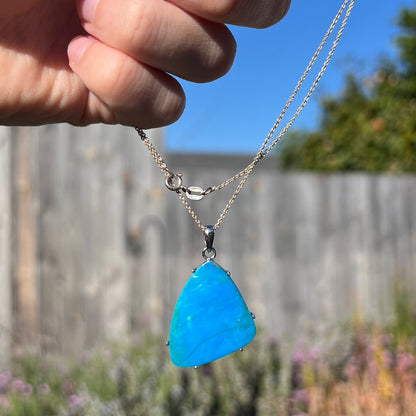 A Chilean blue opal necklace being dangled by its chain outdoors in natural sunlight.