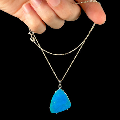 A Chilean blue opal necklace being dangled by its chain in a hand against a black background.