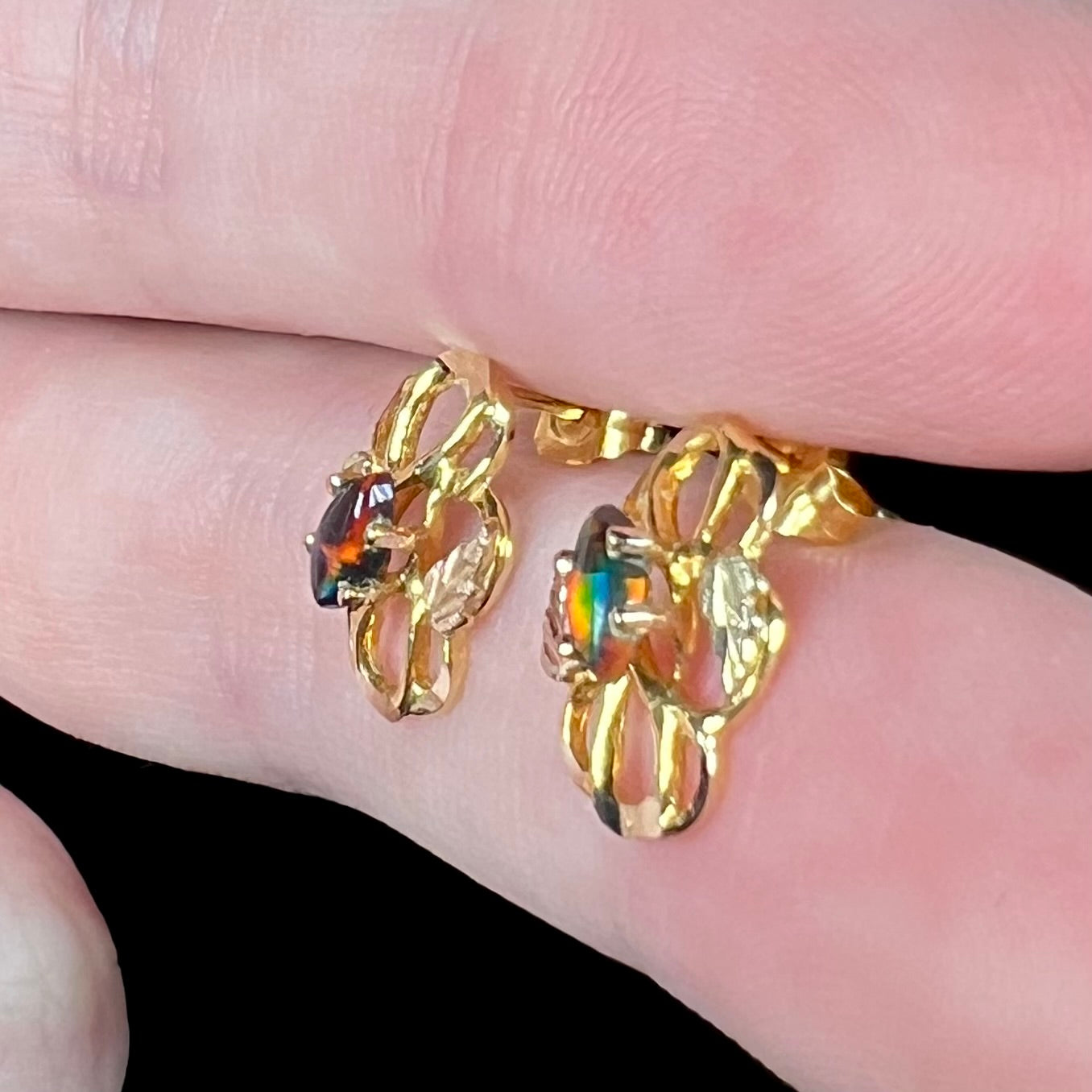 Pair of colorful black opal in Black Hills gold stud earrings, shown close-up between fingers