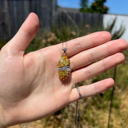 Hand holding an Ethiopian fire opal cluster pendant in the sunlight; the opals show colorful green flashes atop warm, orange body tones.