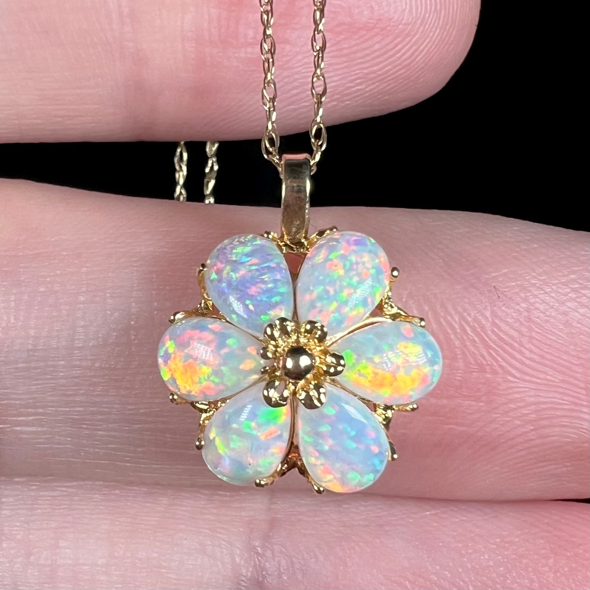 Close-up macro of a colorful lab-created opal flower design pendant in 14k yellow gold, shown held in a hand on a chain