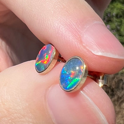 Pair of colorful opal triplet stud earrings in 9k yellow gold bezels, held between two fingers and viewed from angle in sunlight