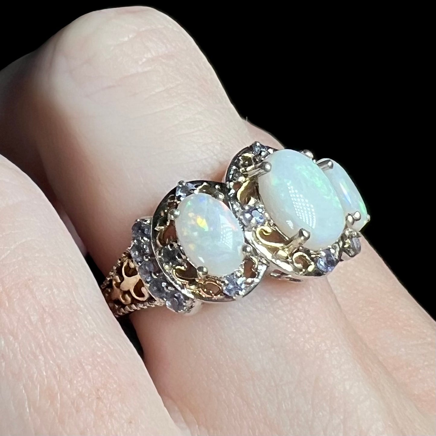 Finger wearing a Coober Pedy, Australian opal and tanzanite ring made from sterling silver with gold-tone open filigree details.