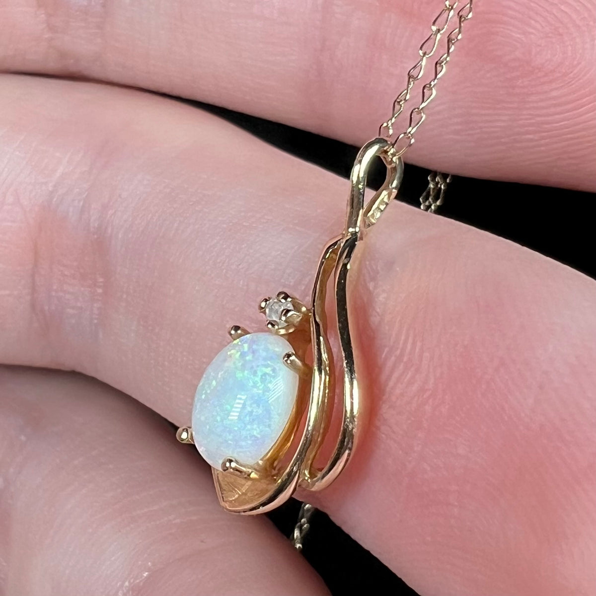 Side close-up view of Coober Pedy, Australian opal and diamond pendant in 14k gold with chain, held in hand