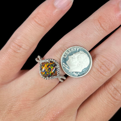 A lab-created black opal solitaire ring with orange color play in sterling silver, shown on a hand next to a dime coin.