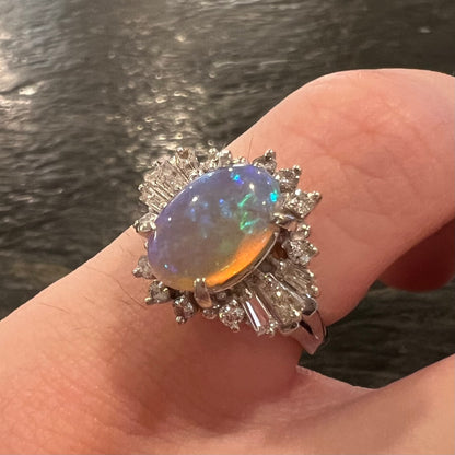 A vintage crystal opal and diamond ballerina style ring shown on pinky finger in dimly-lit environment; the opal has subtle purplish and green colors