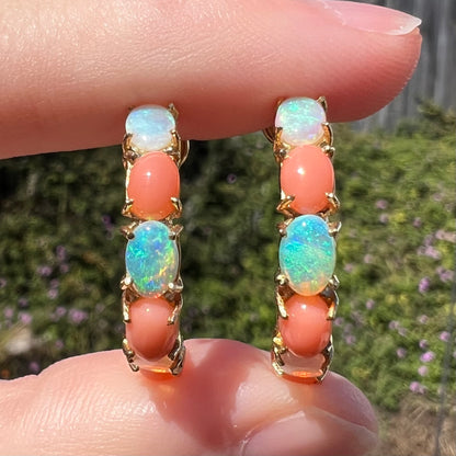 Close-up of vintage crystal opal and pink coral semi-hoop earrings, held between two fingers and shown from front view in sunlight