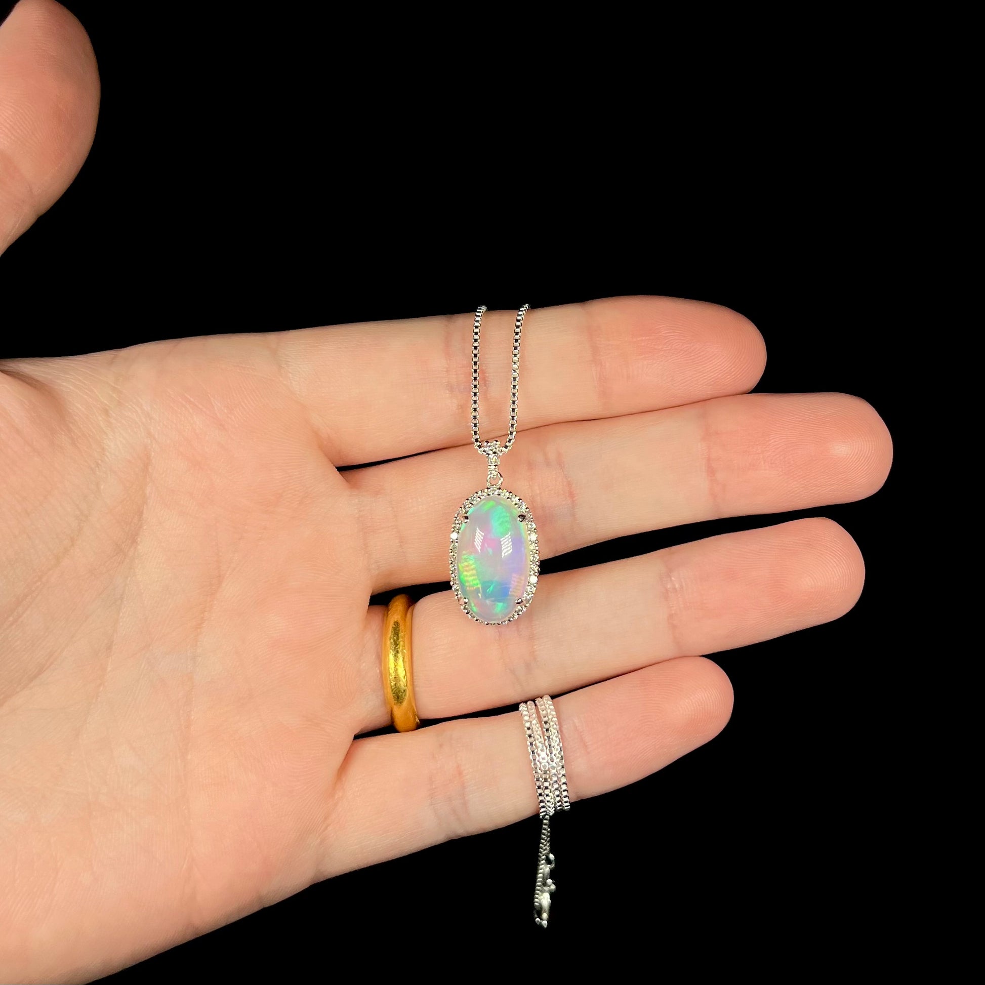 Hand holding a crystal opal and diamond halo necklace made from platinum against a black background under LED studio lights.
