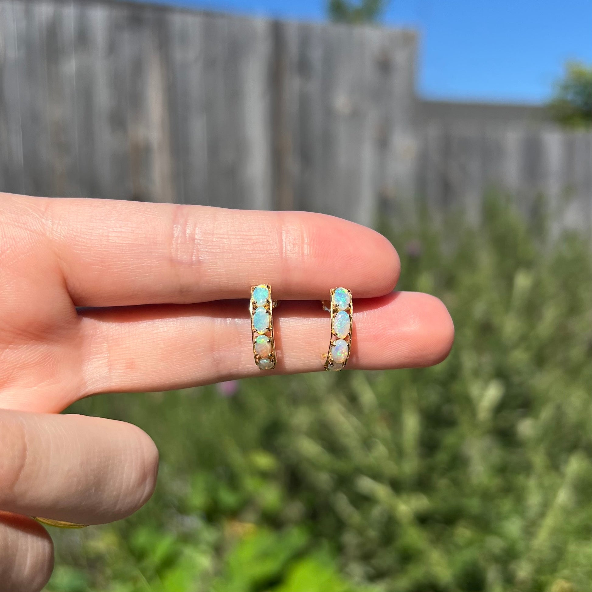 Two fingers holding a pair of yellow gold crystal opal semi-hoop earrings in natural sunlight against a blurred outdoor backdrop.