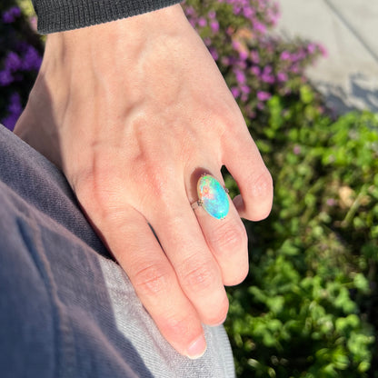 An oval cut crystal opal ring worn on a hand.  The opal shows blue color play.