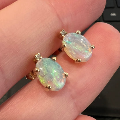 Close-up view of a pair of Australian crystal opal and diamond stud earrings held between two fingers, focused on dark gypsum inclusions.