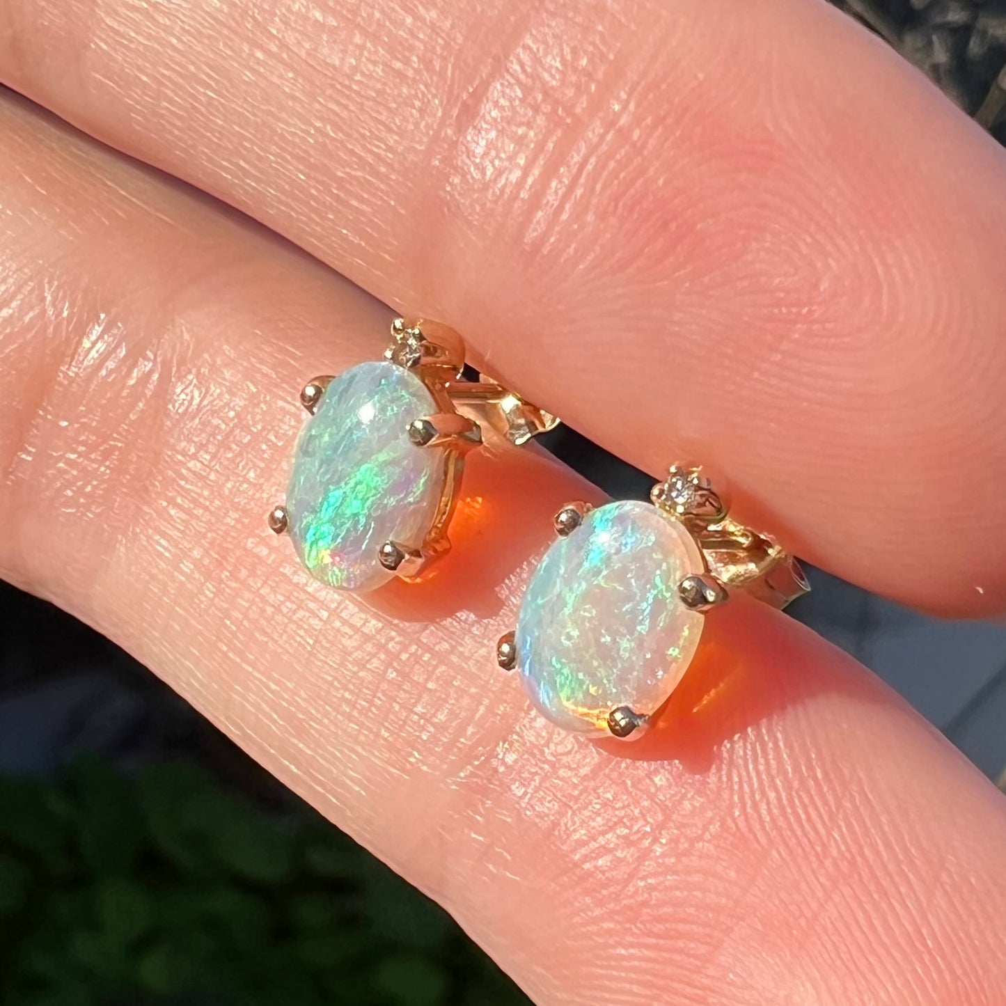 A pair of Australian crystal opal and diamond stud earrings in gold with green-blue color play shown close-up from an angle in the sunlight.