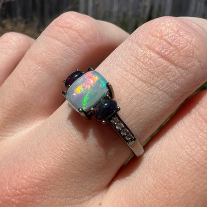 Angled close-up of cushion and oval cut three-stone Ethiopian opal ring with CZ accents and black rhodium finish, worn on finger in natural light