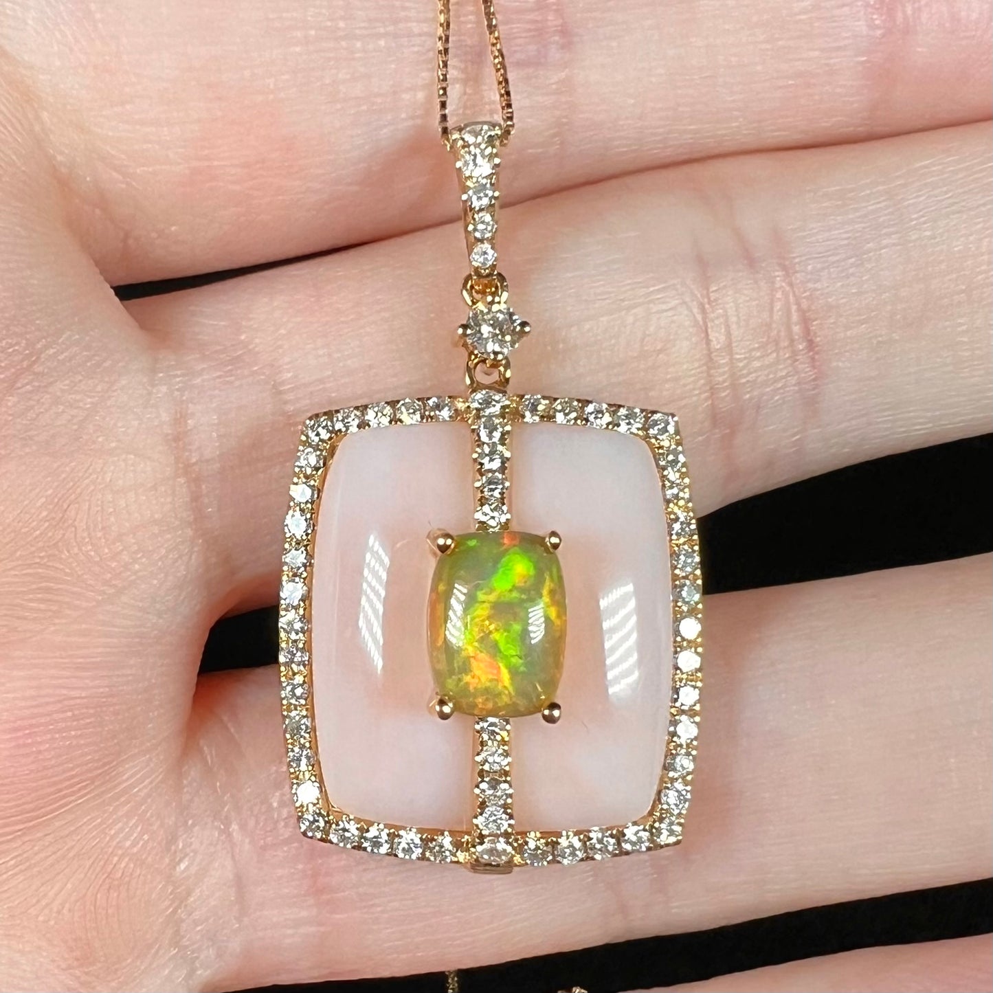 Macro view of a cushion cut Ethiopian fire opal and Peruvian pink opal necklace with diamond accents in yellow gold, held in a hand.