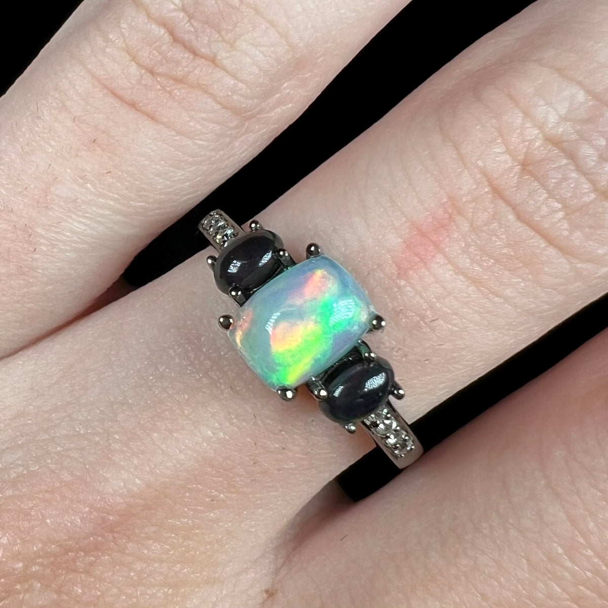 Close-up of cushion cut Ethiopian opal ring with two smoked black opal accents in black rhodium finish, worn on finger