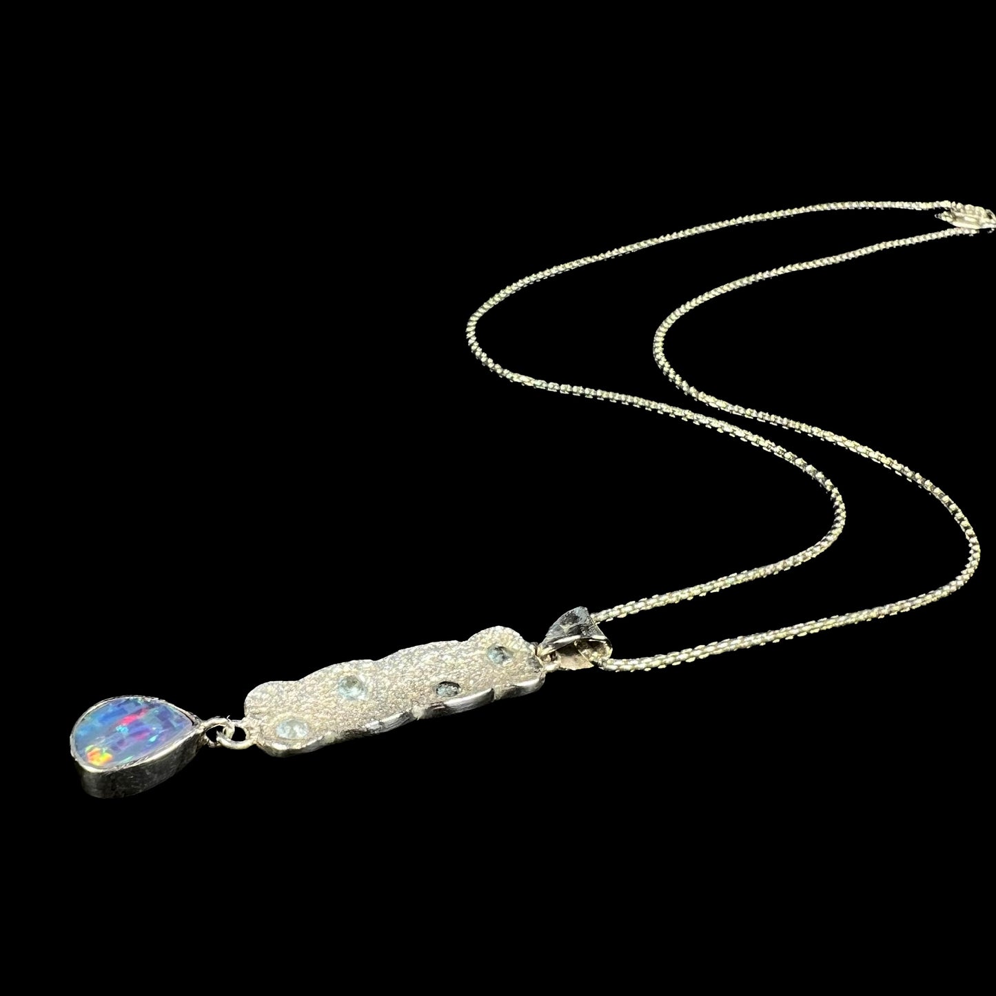 A custom-made sterling silver opal doublet and blue topaz drop necklace shown laying against a black background, its chain flowing.