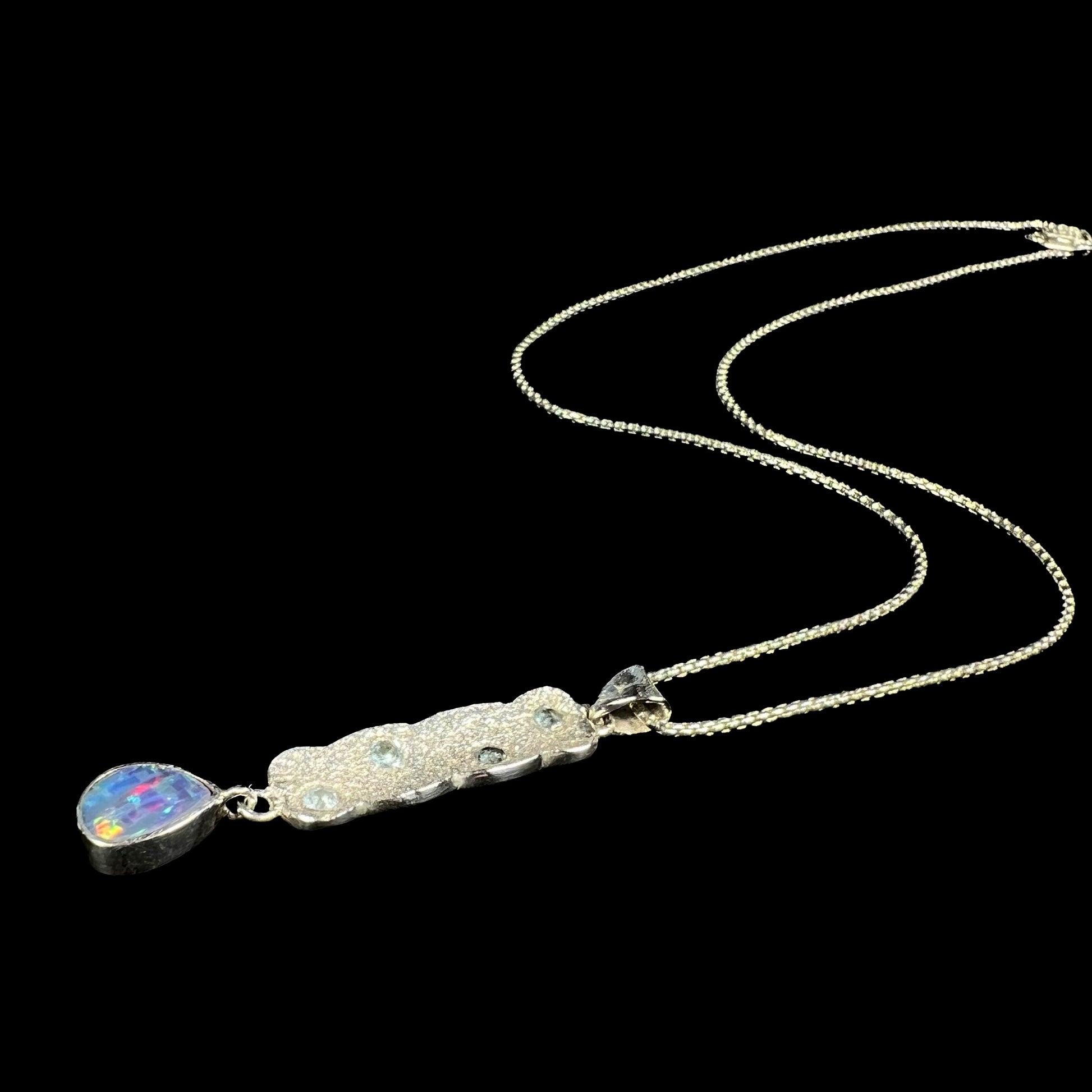 A custom-made sterling silver opal doublet and blue topaz drop necklace shown laying against a black background, its chain flowing.