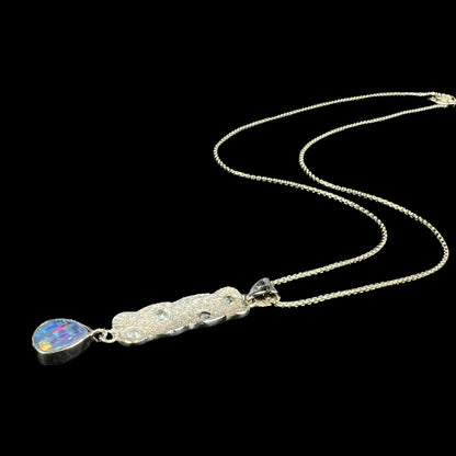 A custom-made sterling silver opal doublet and blue topaz drop necklace shown laying against a black background, its chain flowing.