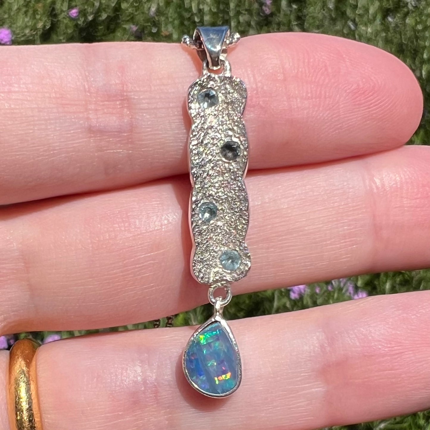 Close-up of a custom-made Australian opal doublet pendant with a textured sterling silver design and blue topaz accents, shown in natural light.