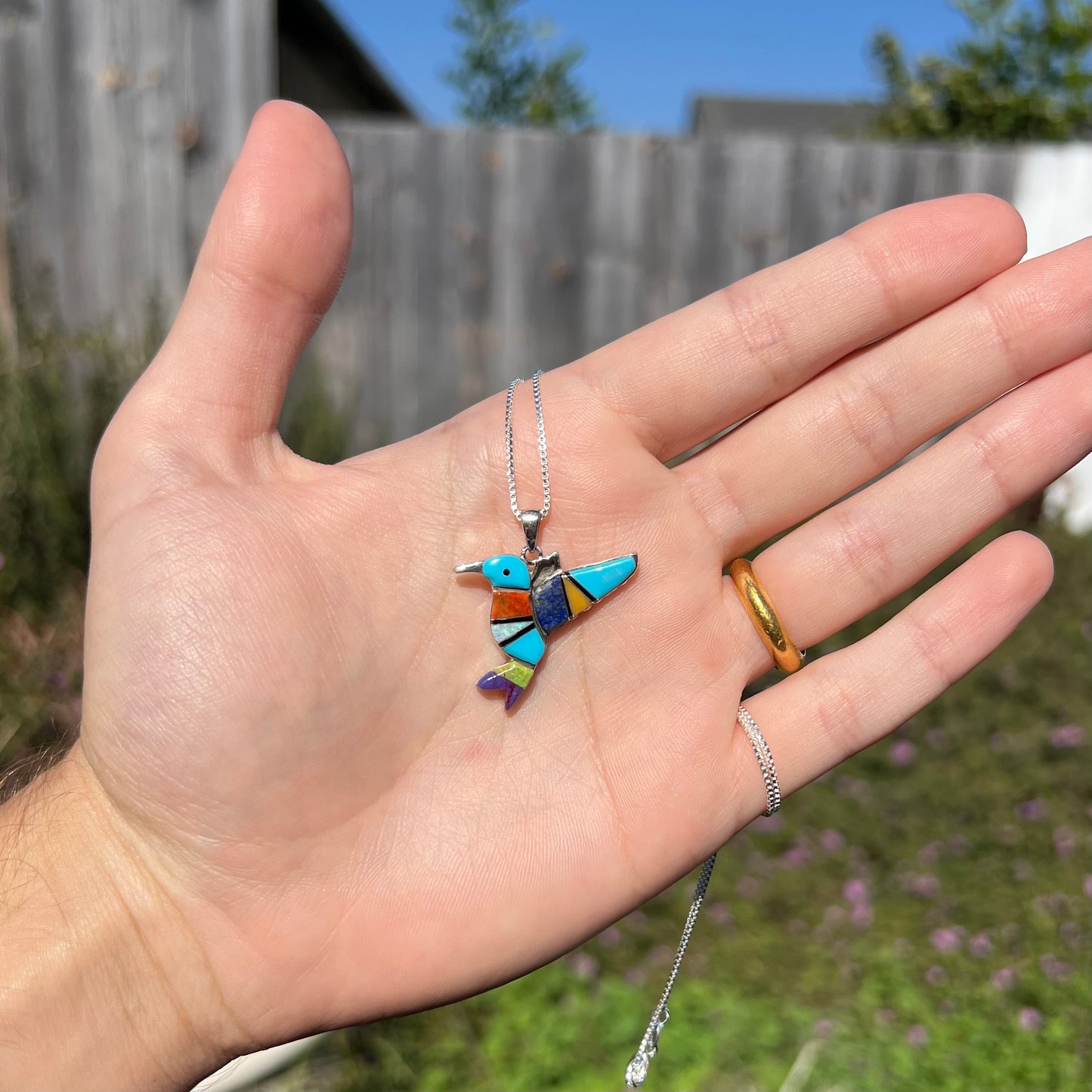 A multi-stone inlay hummingbird necklace in sterling silver, shown in a hand in the sunlight.