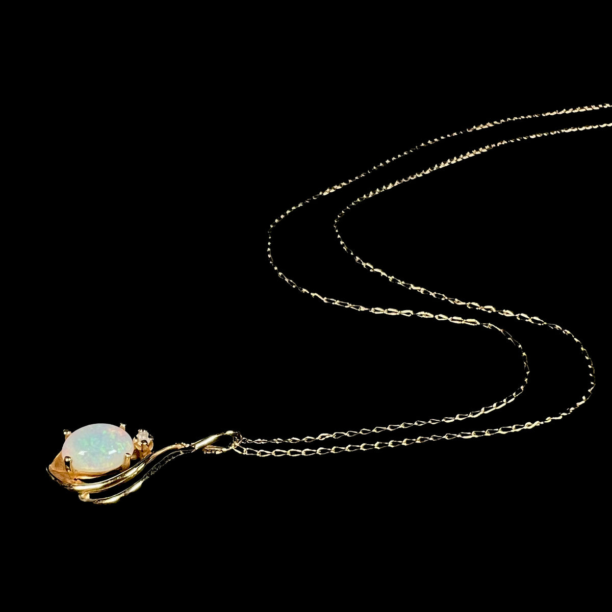 Dainty 14k gold women's opal and diamond necklace shown from angle laying on black background, the chain flowing behind the pendant