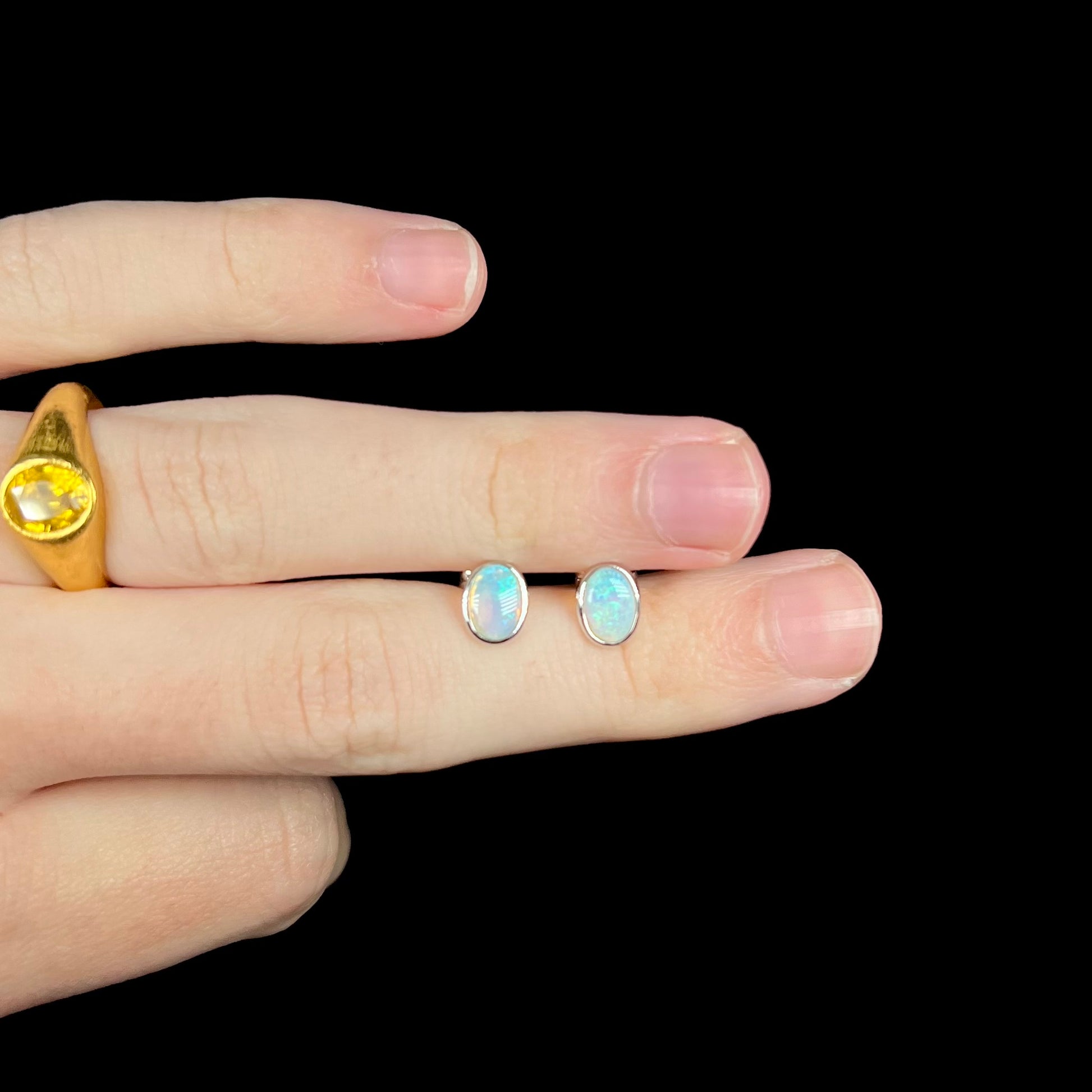 Two dainty Australian crystal opal stud earrings held between fingers of a hand under LED light.