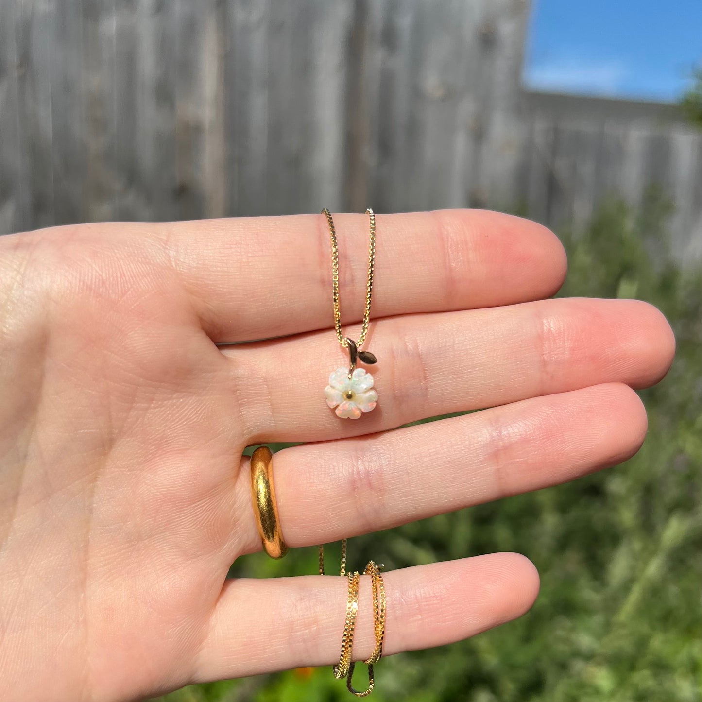 Hand holding a dainty Australian white opal flower pendant on a box chain in natural sunlight against blurred outdoor background.