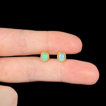Dainty bezel set opal stud earrings with green flashes of color held between two finger under LED lighting against black background.