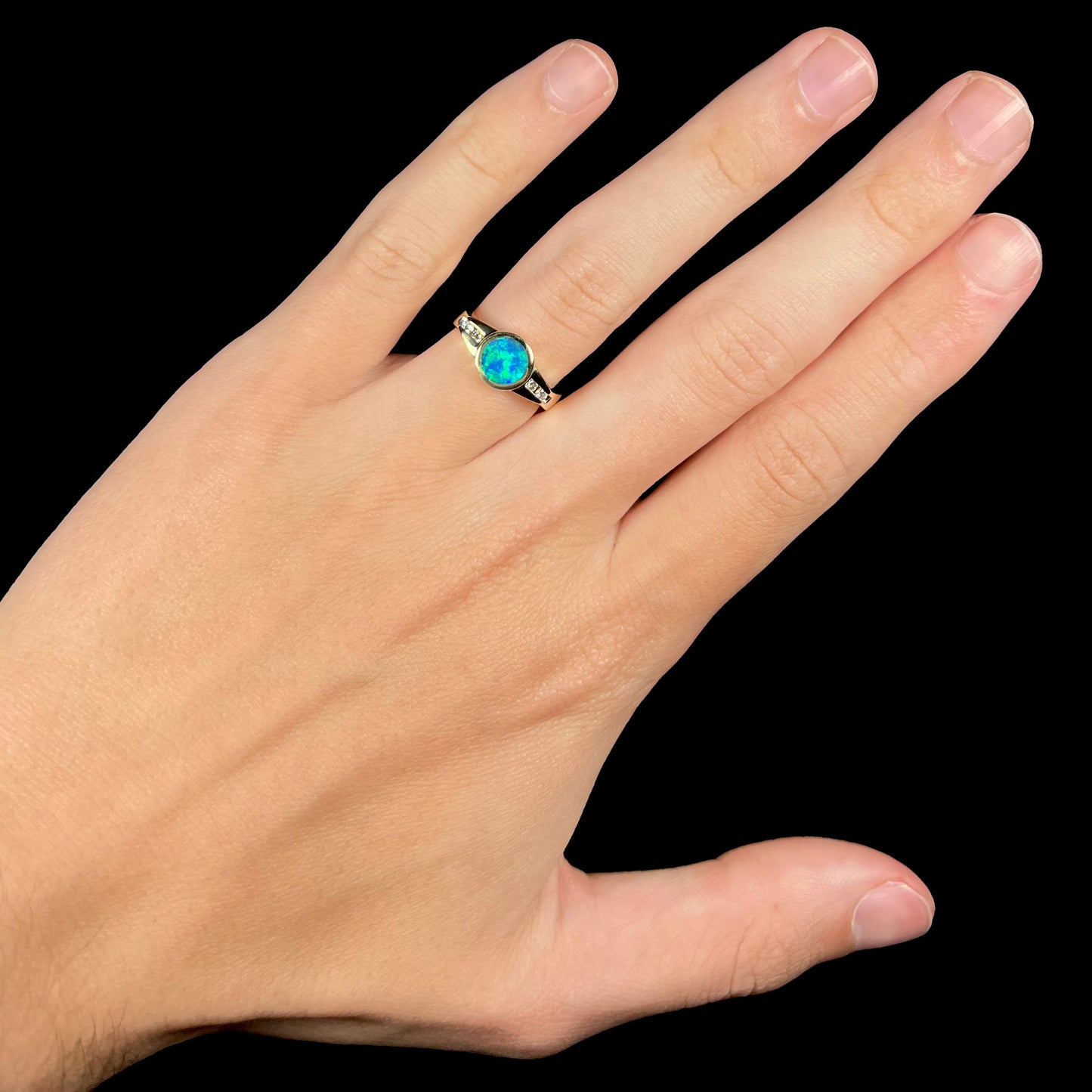Hand wearing a round, dainty black boulder opal and diamond ring against a black backdrop.  Studio light shows green flashes in the opal.