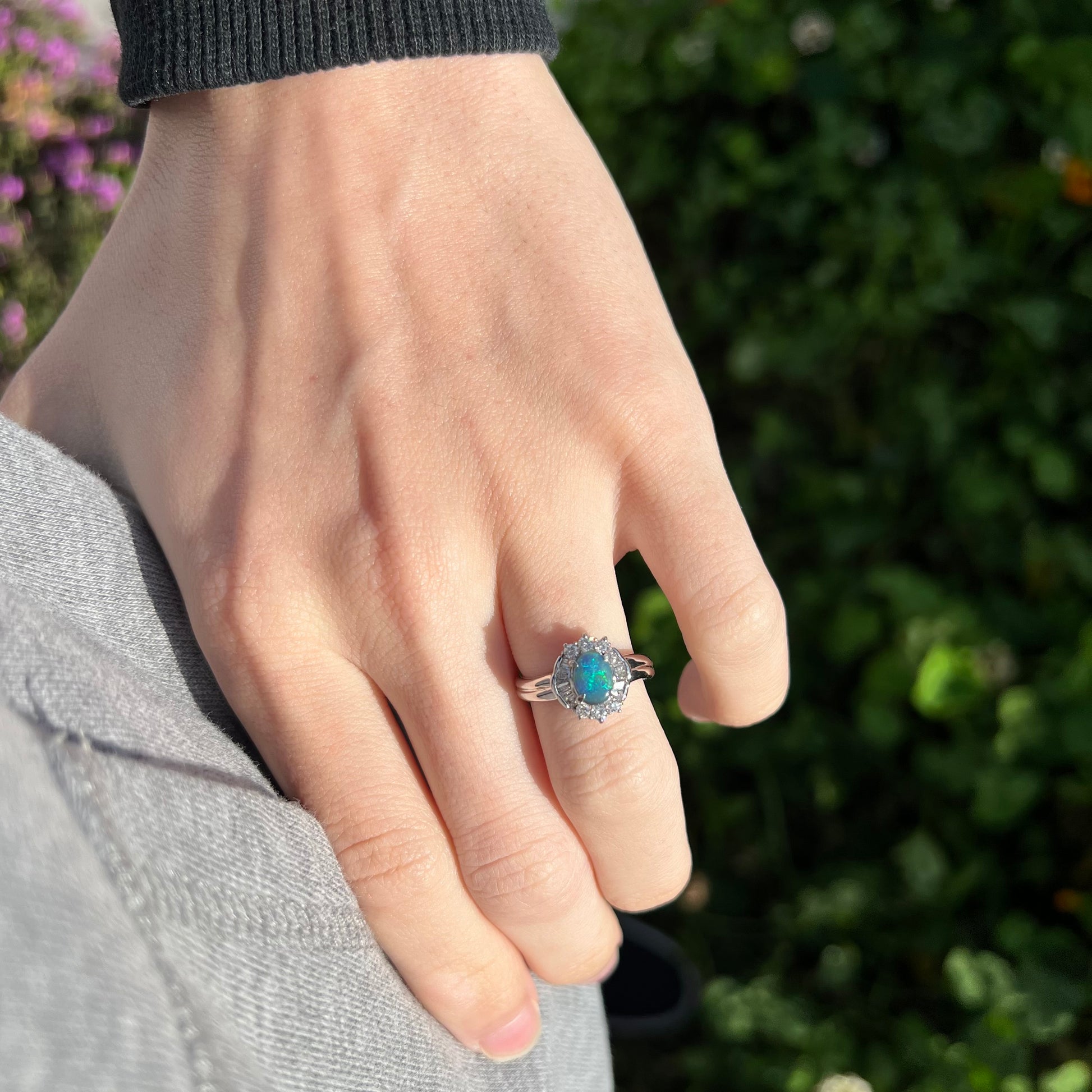 A dainty black opal and diamond halo engagement ring worn on a hand in the sunlight.  Green bushes are in the background.