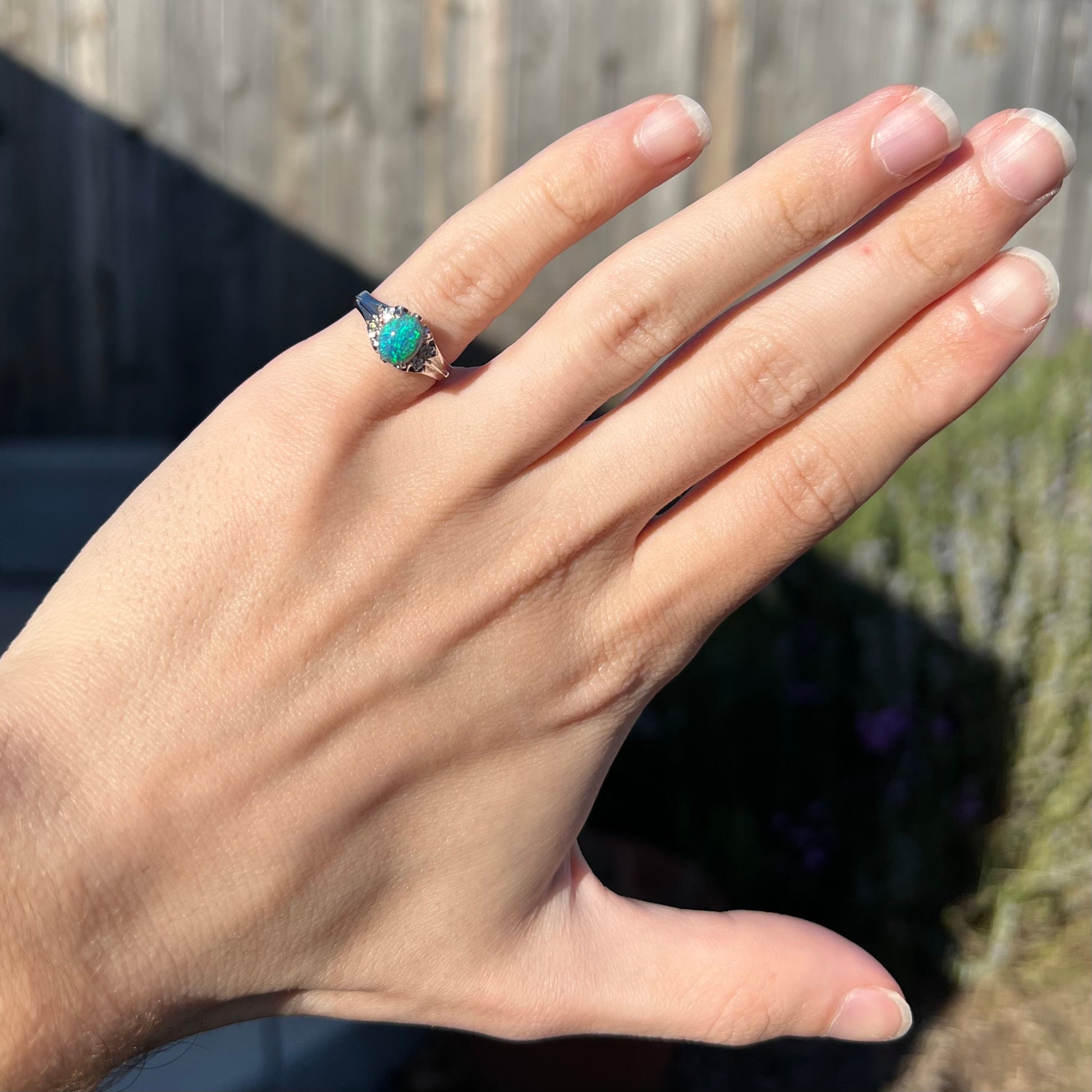 A dainty black opal and diamond platinum ring worn on a hand in the sunlight.