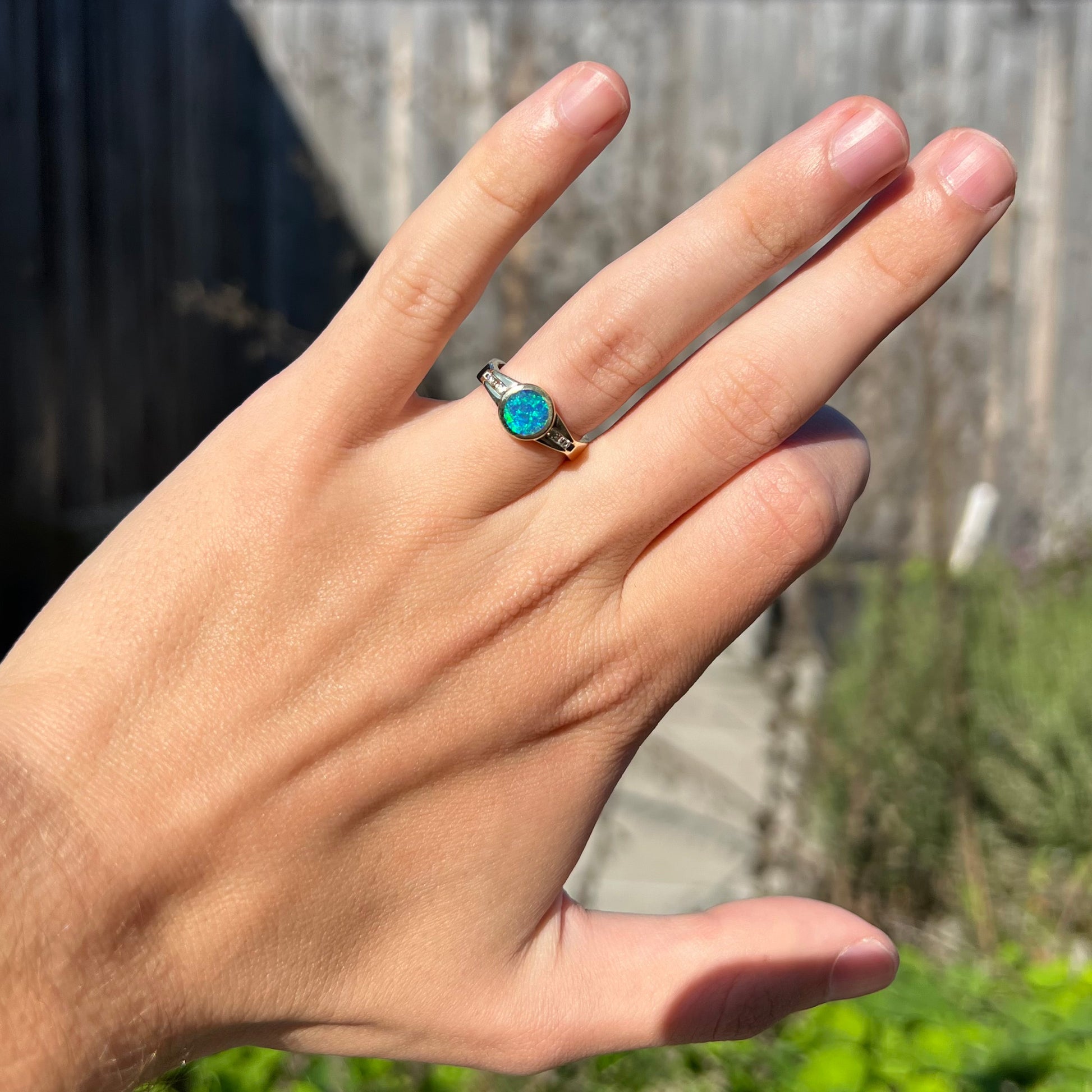 A dainty boulder opal engagement ring with a deep blue body and gentle green fire, worn on a hand in the sunlight against a blurred outdoor backdrop.