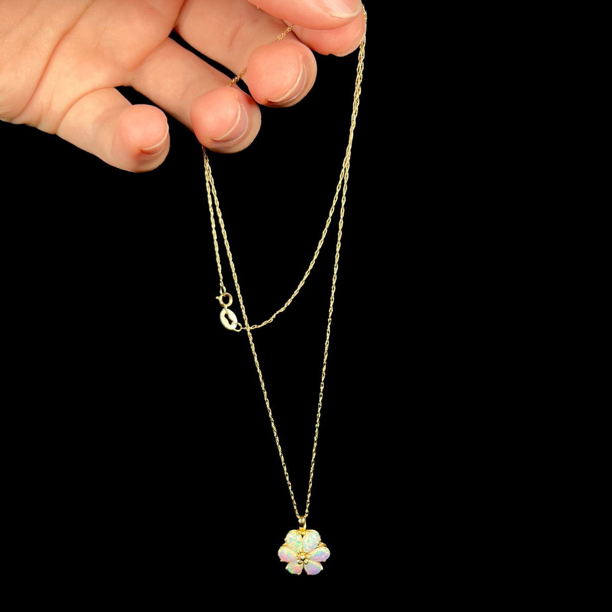 A dainty 14k gold flower necklace dangled by its chain from a hand; the pendant is set with a cluster of six lab-created opals