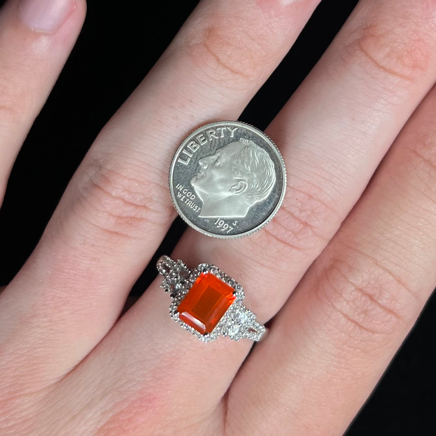 Hand wearing a dainty emerald cut fire opal ring.  A US dime coin rests on the hand next to the ring for scale.