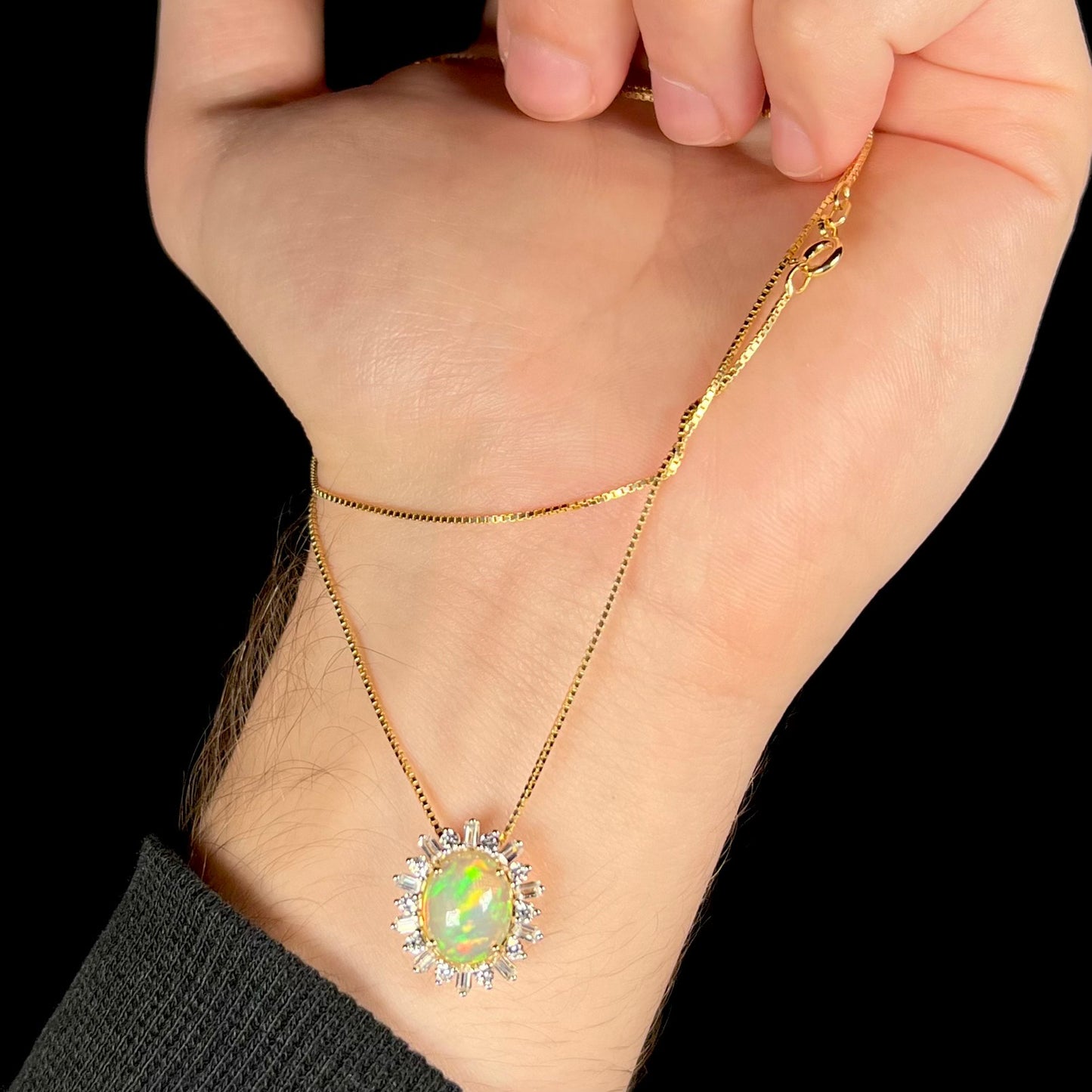 Hand dangling dainty Ethiopian opal halo necklace from its box chain, the 14k gold pendant rests on person's wrist.